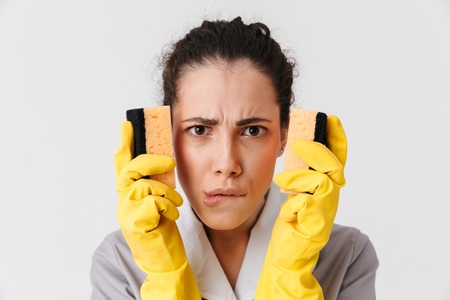 Portrait of a confident young housemaid dressed in uniform and rubber gloves holding sponges isolated over white backgroundの写真素材