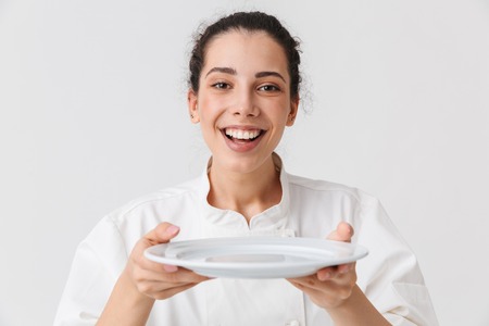 Portrait of a cheerful young woman cook dressed in uniform showing empty plate isolated over white backgroundの写真素材