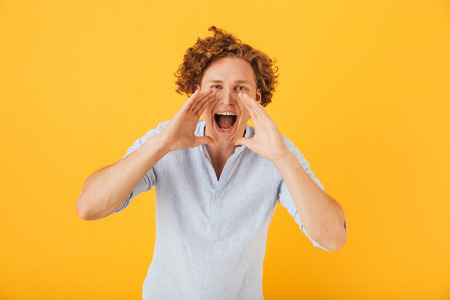 Photo of caucasian curly man 20s shouting or calling while holding hands at face isolated over...