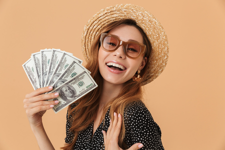 Image of charming european woman 20s wearing straw hat and sunglasses smiling and holding fan of dollar banknotes isolated over beige backgroundの写真素材
