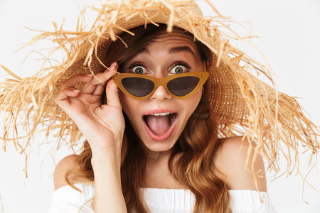 Portrait closeup of excited adorable woman 20s wearing big straw hat looking at camera from under sunglasses isolated over white backgroundの写真素材