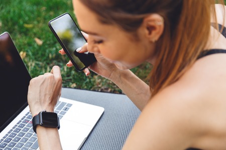 Close up of a smiling fitness girl synchronizing her smart watch and mobile phone while laying on a...