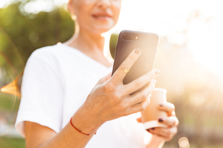 Cropped blurry image of european woman wearing casual clothing holding cell phone and takeaway coffee in hands during walk outdoorの写真素材