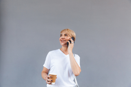 Picture of happy adult woman wearing white t-shirt standing isolated over gray wall and holding coffee from paper cup while talking on mobile phoneの写真素材