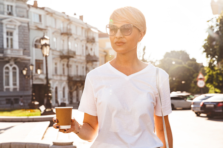 Image of caucasian blond woman wearing white t-shirt and sunglasses walking through city street in summer and drinking takeaway coffee from paper cupの写真素材