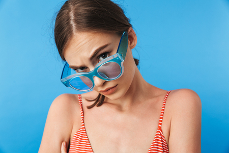 Portrait of an anry young girl in swimsuit wearing sunglasses looking at camera isolated over blue backgroundの写真素材