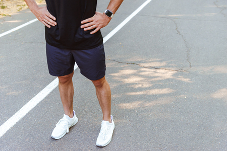 Cropped image of a young sportsman with smartwatch standing with arms on hips on a roadの写真素材
