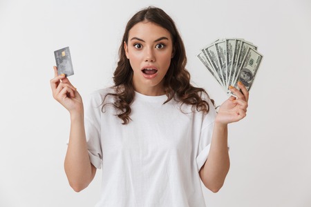 Portrait of a shocked young casual brunette woman holding credit card and money banknotes isolated over white backgroundの写真素材