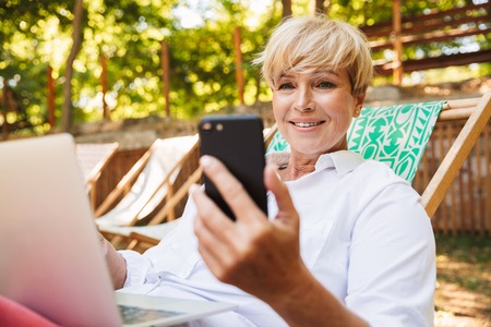 Smiling mature woman holding mobile phone while resting at the park with laptop computerの写真素材