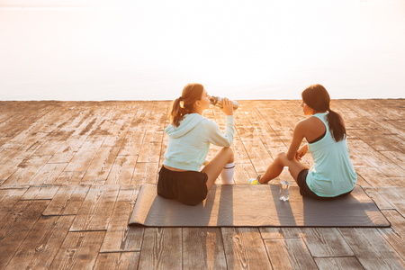 Back view picture of amazing young two sports women friends outdoors on the beach sitting talking with each otherの写真素材