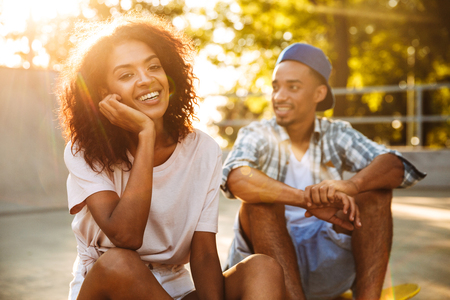 Portrait of a laughing young african couple with skateboard sitting together at the skate parkの写真素材