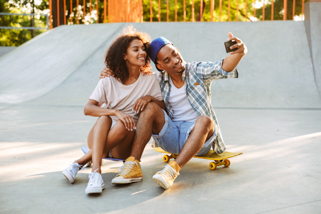 Portrait of a happy young african couple with skateboards taking a selfie together at the skate parkの写真素材