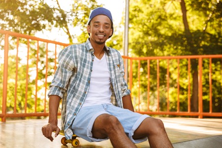 Smiling young african man with skateboard sitting on a ramp at the skate parkの写真素材