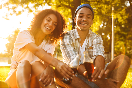 Image of cute cheerful young african loving couple sitting outdoors in park drinking soda.の写真素材