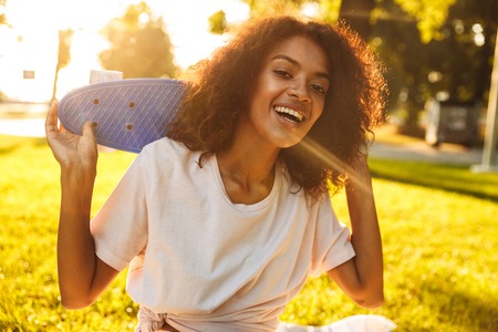 Delighted young african girl holding skateboard while sitting at the parkの写真素材