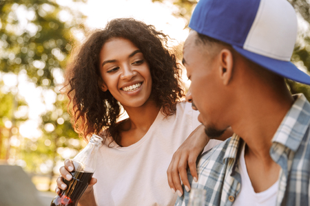 Portrait of a cheerful young african couple holding bottles with fizzy drink sitting together at the skate parkの写真素材