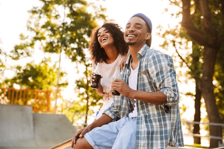 Portrait of a laughing young african couple holding bottles with fizzy drink while sitting together at the skate parkの写真素材