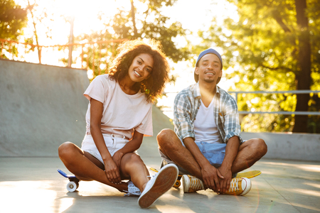 Portrait of a smiling young african couple with skateboard sitting together at the skate parkの写真素材