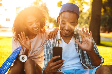 Portrait of a happy young african couple with skateboards taking a selfie together at the park, wavingの写真素材