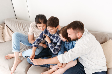 Image from above of cheerful family with two children sitting on sofa together at home and taking selfie photo on mobile phoneの写真素材