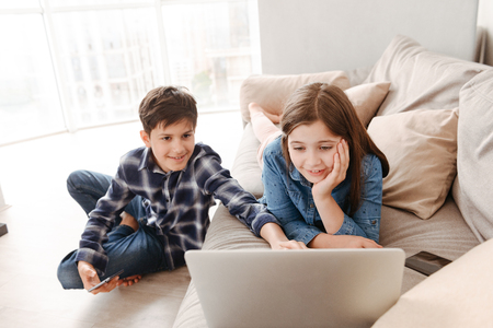 Photo of two teen child girl and boy 8-10 resting on sofa at home while using smartphone and laptopの写真素材