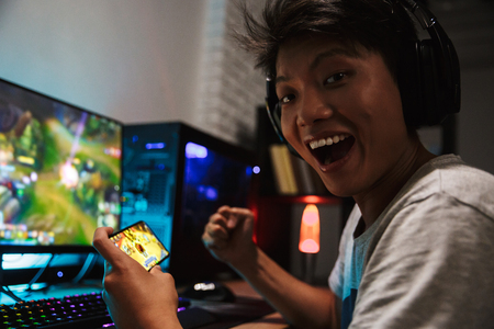 Asian happy gamer boy rejoicing while playing video games on smartphone and computer in dark room wearing headphones and using backlit colorful keyboardの写真素材