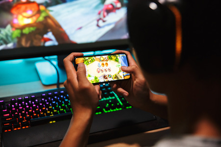 Teenage gamer boy playing video games on smartphone and computer in dark room wearing headphones and using backlit colorful keyboard