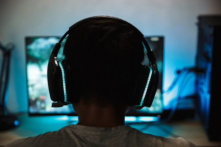 Back view closeup of young gamer boy playing video games online on computer in dark room wearing headphones with microphoneの写真素材