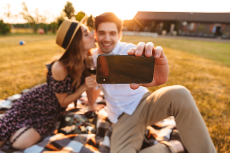Photo of young cute loving couple sitting by dating outdors on picnic holding glasses of wine take a selfie by mobile phone.の写真素材