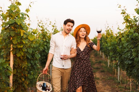 Photo of young cute happy loving couple outdoors drinking wine holding basket with bottles.
