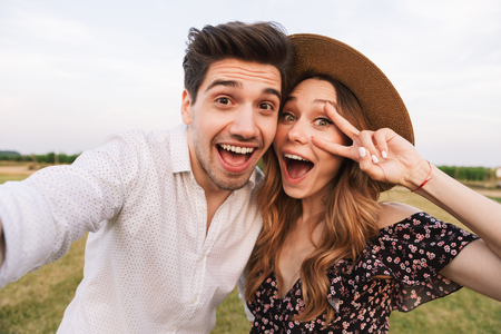 Picture of young happy cute loving couple outdoors hugging with each other take a selfie by camera with peace gesture.の写真素材