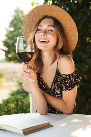 Image of cute pretty young woman sitting in cafe outdors in park with book holding glass drinking wine.の写真素材
