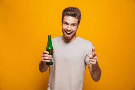 Image of handsome man standing isolated over yellow wall backgroung drinking beer pointing to you looking at camera.の写真素材