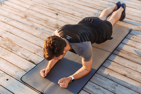 Photo from above of adult european man 20s in tracksuit lying on mat and doing plank while working out on wooden pier at seaside in morningの写真素材