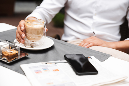 Close up of a man drinking coffee while sitting at the cafe table outdoors with blank screen mobile phoneの写真素材