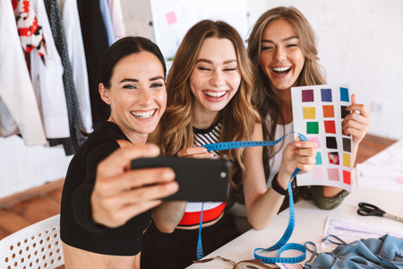 Three happy young women clothes designers working together at the atelier, sitting at the table and taking selfieの写真素材
