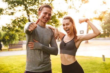 Image of happy excited fitness sport loving couple friends in park outdoors showing biceps and thumbs up gesture looking camera.の写真素材