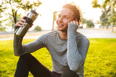Picture of handsome fitness sportsman in park outdoors holding bottle with water.