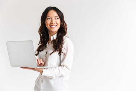 Smiling young asian businesswoman standing with laptop computer over white background, looking awayの写真素材