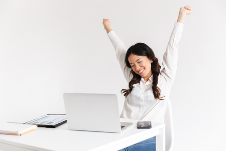Photo of successful chinese businesslike woman with long dark hair smiling with raised arms while working with documents and laptop isolated over white backgroundの写真素材