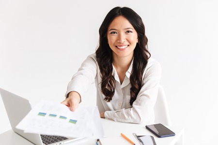 Smiling young asian woman giving paper graph while sitting at the desk with laptop computer isolatedの写真素材