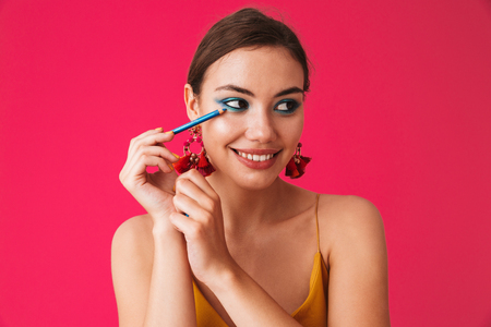 Portrait of gorgeous stylish woman 20s wearing earrings smiling and applying makeup with blue eyeliner isolated over pink backgroundの写真素材