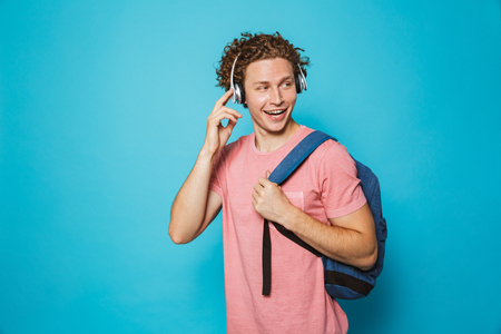 Portrait of attractive man 18-20 with curly hair wearing backpack listening to music via headphones isolated over blue backgroundの写真素材