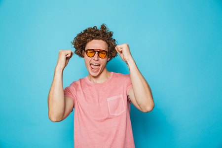 Portrait of fashion guy 20s with brown curly hair wearing modern glasses screaming and rejoicing isolated over blue backgroundの写真素材