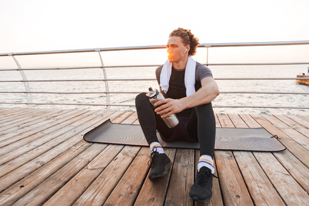 Photo of handsome strong young sportsman outdoors at the beach sitting listening music with earphones drinking water.の写真素材