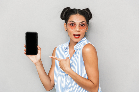Portrait of an excited young woman in sunglasses isolated, showing blank screen mobile phoneの写真素材