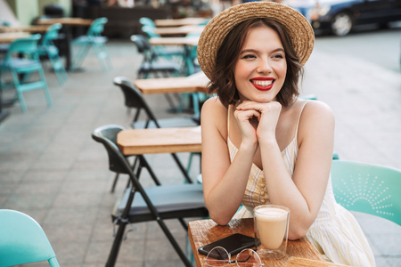 Smiling woman in dress and straw hat drinking coffee while sitting by the table in city cafeの写真素材