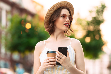 Pretty woman in dress, straw hat and sunglasses posing with cup of coffee and smartphone while looking away outdoorsの写真素材