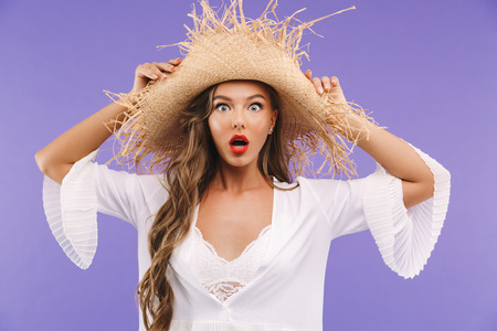 Photo of young excited woman 20s wearing dress and straw hat looking at camera with open mouth and big eyes isolated over violet backgroundの写真素材