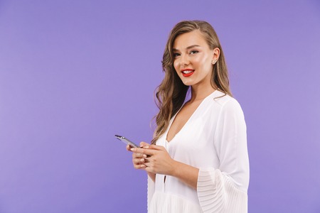Portrait of a smiling young woman in white dress standing isolated, using mobile phone